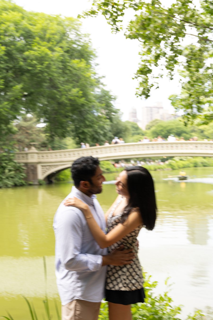 Akshay and Daniela embracing by the lake with Bow Bridge behind them